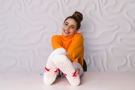 The portrait of a beautiful brunette with her hair tied up in a bun, wearing an orange turtleneck and deer socks, has thrown her legs up on the table and happily poses in a pastel pink room.の写真素材