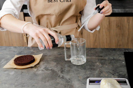 Close up of A young pastry chef in a beige apron pours syrup in to cup for application onto a...