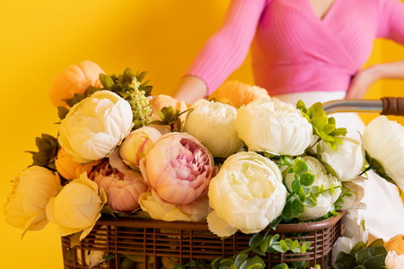 A charming, bright and happy girl in a pink top and short skirt riding a bike with a basket filled with flowers on an isolated yellow background. Close-up.の写真素材