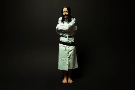 A mentally unstable and ill woman in a straitjacket with a cloth tied over her mouth in a psychiatric hospital on a black isolated background. Mental disorders concept. Crazy preson.の写真素材