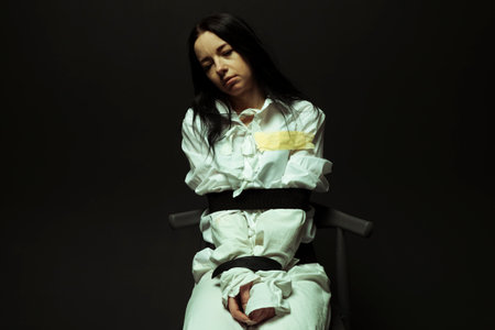 A mentally unstable and ill woman in a straitjacket sitting in a chair in a psychiatric hospital on a black isolated background. Mental disorders concept. Crazy preson.の写真素材