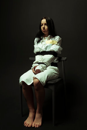 A mentally unstable and ill woman in a straitjacket sitting in a chair in a psychiatric hospital on a black isolated background. Mental disorders concept. Crazy preson.の写真素材