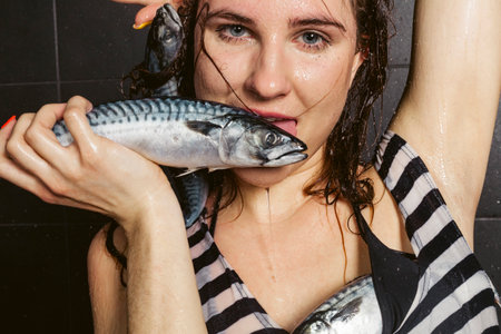A young brunette girl in a long striped wet shirt holds and poses with a mackerel or fish while taking a shower. Black matt ceramic tiles in the background. wet skin and hair.の写真素材
