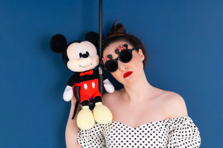 A woman with bright red painted lips, dressed in a black and white polka dot dress and a black round sunglasses with plush toy, isolated on blue. 50s style.の写真素材