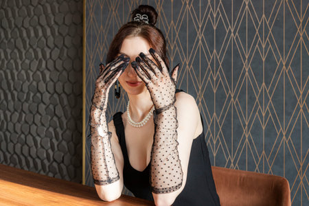 A beautiful, gorgeous and elegant girl in a black dress and Mesh Sheer gloves on her hands, covering her eyes. formal or upscale event.の写真素材