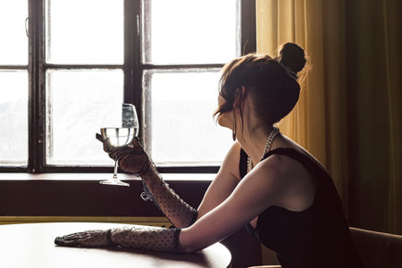 Beautiful, gorgeous and elegant girl in a black dress and Mesh Sheer gloves on her hands, sitting at a table with a glass in her hand. Concept of elegance and sophistication. Formal or upscale event.の写真素材