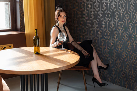 Beautiful, gorgeous and elegant girl in a black dress and Mesh Sheer gloves on her hands, sitting at a table with a glass in her hand. Concept of elegance and sophistication. Formal or upscale event.の写真素材
