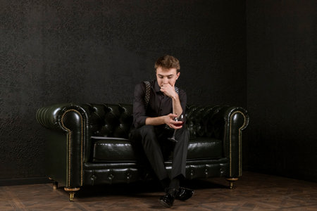 A young man is sitting on an elegant couch in dark room with a glass of wine in his hand and music sheets and contemplating a composition. The process of creation.の写真素材