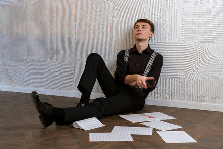 Elegant, handsome young man in a black suit and suspenders is sitting on a floor with a music sheets...