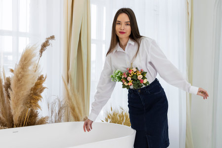 Portrait of a young beautiful brown-eyed girl in a white shirt, with flowers in her hands, posing in a minimalist modern bathroom. Beauty and Freshness concept.の写真素材