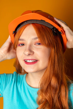 A red-haired girl wearing a construction helmet is showing a thumbs-up on an isolated orange background. Emphasizing safety regulations.の写真素材