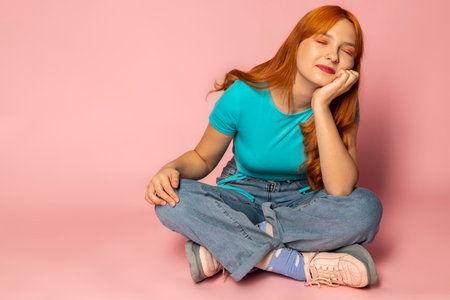 A happy red-haired young girl sitting and dreaming against an isolated pink background.の写真素材
