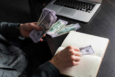A young serious man in a gray formal suit and glasses sits at his desk and counts cash. A...