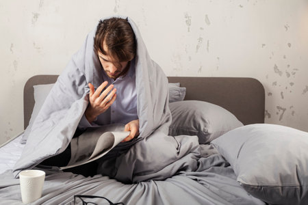 An upset young man is sitting on a bed, wrapped in a blanket, and reading bad news from a newspaper.の写真素材