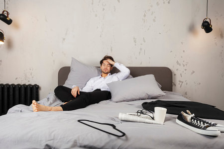 A Handsome young man in dressy clothes lying on a bed with headache, a mess around him.
