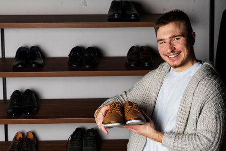 A young man holding a pair of fancy leather shoes. Shelf with boots on the background.の写真素材