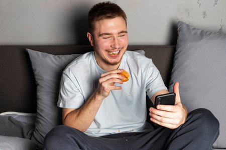 A young man is sitting on the bed and eating, while talking on a video call on his phone or watching...