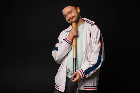 A young, tough but smiling guy from the streets dressed in a tracksuit holding a baseball bat, posing, isolated on a black background. Urban street culture, Street gang, urban fashion.の写真素材