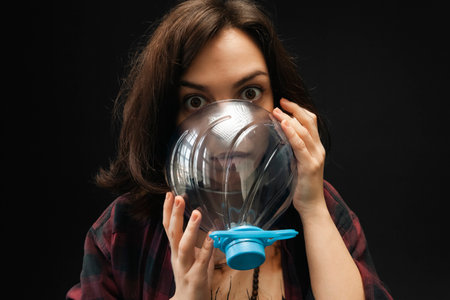 The portrait of a girl with big stunned eyes is breathing through half of a cut plastic bottle. Isolated on a black background. Concept of air and plastic pollution.の写真素材