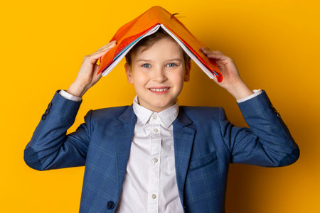 A smiling little boy in a blue suit and white shirt, neatly dressed, covers his head with a textbook. Isolated on a yellow background.の写真素材