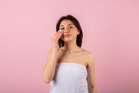 An attractive teenage girl wrapped only in a white towel is happily smiling and using a cotton round or pad. isolated on a pink background.の写真素材