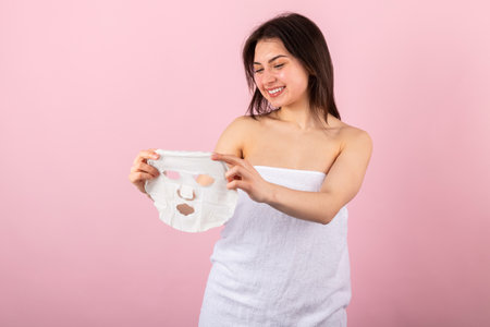 Attractive teenage girl wrapped only in a white towel happily smiling and removing a pre-made facial sheet mask. isolated on a pink background.の写真素材
