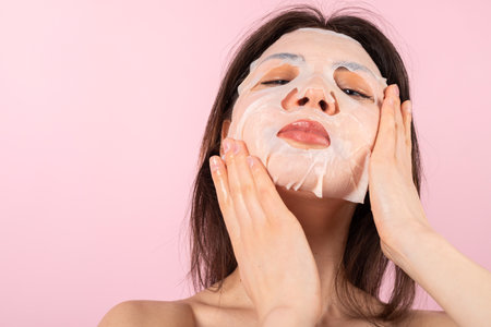 Attractive teenage girl wrapped only in a white towel putting a pre-made facial sheet mask. isolated on a pink background. Close-up.の写真素材