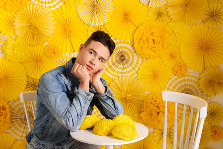 Portrait of a handsome young man in denim clothes posing against a bright, creative room decorated with yellow origami or hand made paper decorations.の写真素材
