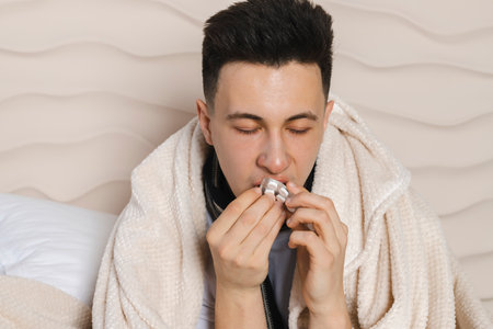 The young man is lying in bed wrapped in a blanket and is sick with a cold, with a red face from the illness. Feeling unwell, flu, fever. taking medicine or pills.の写真素材