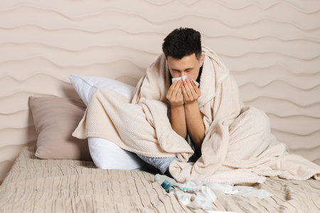 The young man is lying in bed wrapped in a blanket and is sick with a cold, with a red face from the illness. Feeling unwell, flu, fever. blowing his nose, sniffling.の写真素材