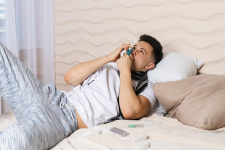 The young man is lying in bed wrapped in a scarf and is sick with a cold, with a red face from the illness. Feeling unwell, flu, fever. asthma, metered-dose aerosol inhaler.の写真素材