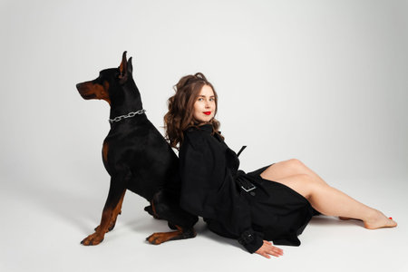 A young beautiful Doberman with a chain around his neck and his owner, elegant girl in a long black coat and bright red lipstick. Isolated on a white background.の写真素材