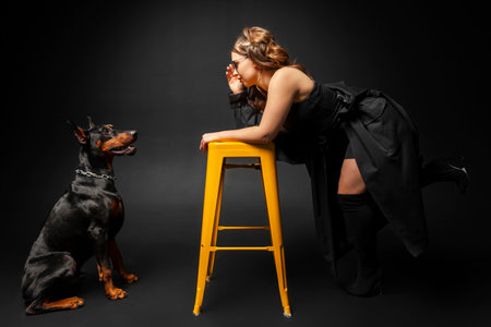 A young beautiful Doberman with a chain on its neck and Stylish girl in a long black coat and bright red lipstick looking at each other. Isolated on a black background.の写真素材