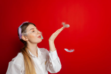 Portrait of a lovely tender girl wearing a feather decorations and blows away feathers from her hands. Isolated on a red background.の写真素材
