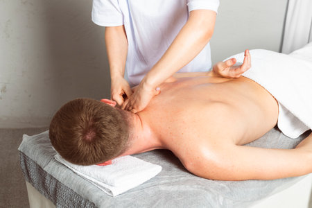 A professional masseuse or massage therapist is massaging the client's shoulder blades while the client lies partially covered with a towel on the massage table. Concept of relaxation and self-care.の写真素材