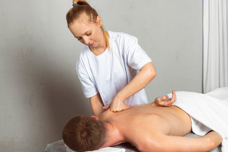 A professional masseuse or massage therapist is massaging the client's shoulder blades while the client lies partially covered with a towel on the massage table. Concept of relaxation and self-care.の写真素材