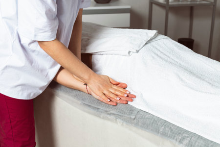 A professional masseuse or massage therapist is massaging the client's hands while the client lies partially covered with a towel on the massage table. Hand to hand.の写真素材