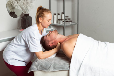 A professional masseuse or massage therapist is massaging the client's neck and shoulders. the client lies covered with a towel on the massage table. profile view.の写真素材