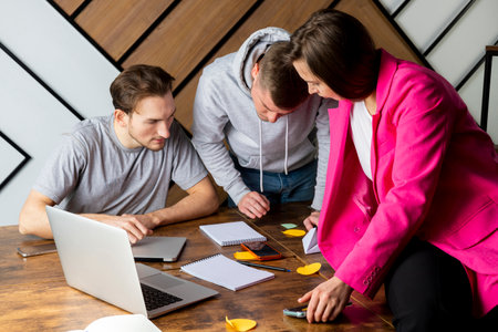 A team of three people is working in the office, communicating and discussing work, looking at notes. IT team in the office, two men and a woman. Young serious entrepreneurs discussing business.の写真素材