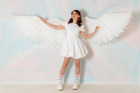 Cute like an angel brunette girl in a fancy short white dress, huge feather wings and bright makeup agains a sky background.の写真素材