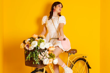 Cute stylish brunette girl, dressed in a fancy short white dress and bright makeup is sitting on a bike with flowers and happily posing and looking away. Isolated on yellow.の写真素材