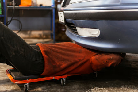 A hardworking mechanic with oil-stained clothing on a mechanics creeper is inspecting a bottom of the car.の写真素材