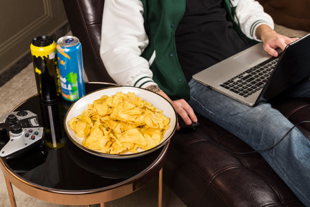 A boy or young man sitting on a couch and playing video games with a laptop while wearing headphones.  A table next to him with chips, soda, and energy drinks. Gamer.の写真素材