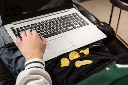 A gamer sitting on a couch and playing video games with a potato chips scattered all around him. An unkempt young man or boy. Dirty hands covered in oil.の写真素材