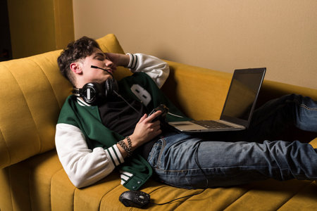 A boy or young man in headphones has fallen asleep on a couch with a controller in his hand and a laptop. The gamer had been playing all night and is now sleeping through the day.の写真素材