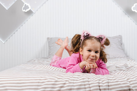 A charming little girl in a pink pajama and robe is lying on her stomach on a bed, posing for a photo.の写真素材