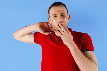 Young handsome healthy man in a red polo shirt and with under eye patches on his face, yawning. Isolated on blue background.の写真素材