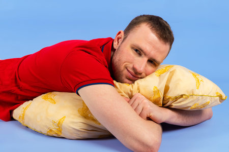 Young handsome healthy man in a red polo shirt is lying on a pillow. Isolated on blue background.の写真素材