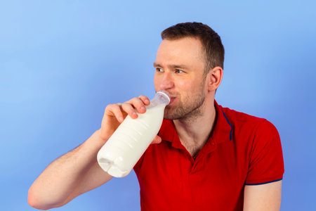 Young handsome healthy man in a red polo shirt is drinking milk from a bottle. Isolated on blue background. full length.の写真素材