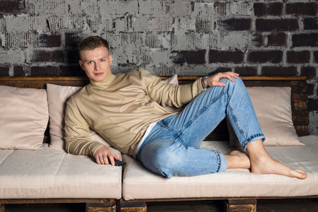 Lazy morning. Man relaxing on comfy sofa at home after finishing work looking aside with glad smile. Calm young guy taking pleasure in awaited weekend enjoying peaceful dreamsの写真素材